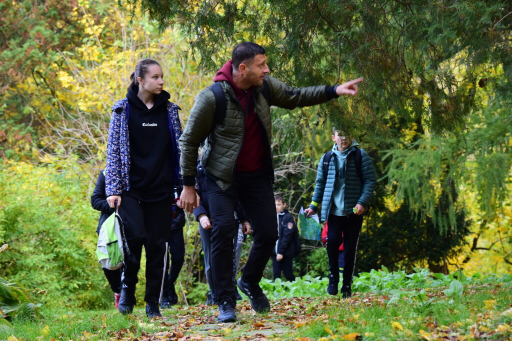 nauczyciel prowadzi uczniów przez park, ręką wskazuje drogę