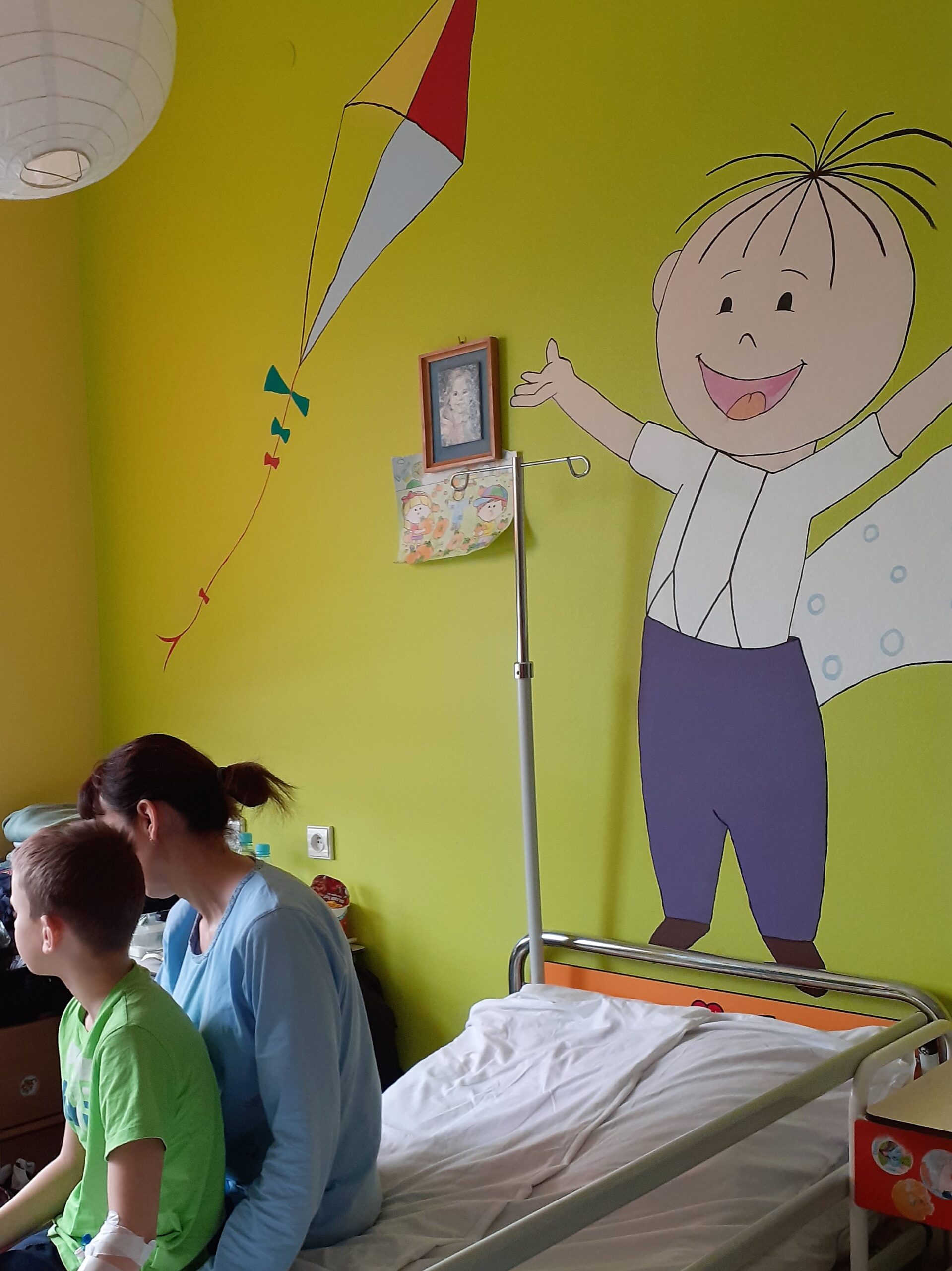 sala szpitalna na pediatrii, na łóżku siedzi kilkuletni chłopiec z mamą, na ścianie postać Lolek z bajki