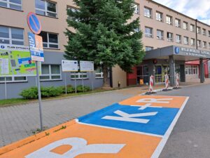 miejsca postojowe kiss and ride, na asfalcie pomalowane na kolor niebieski i pomarańczowy z napisem K+R, znak pionowy z podobnym oznaczeniem, w tle wejście główne do szpitala