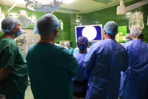 sala operacyjna, szeroki kadr, jeden lekarz prowadzi zabieg, grupa asystentów stoi obok, w tle aparatura medyczna