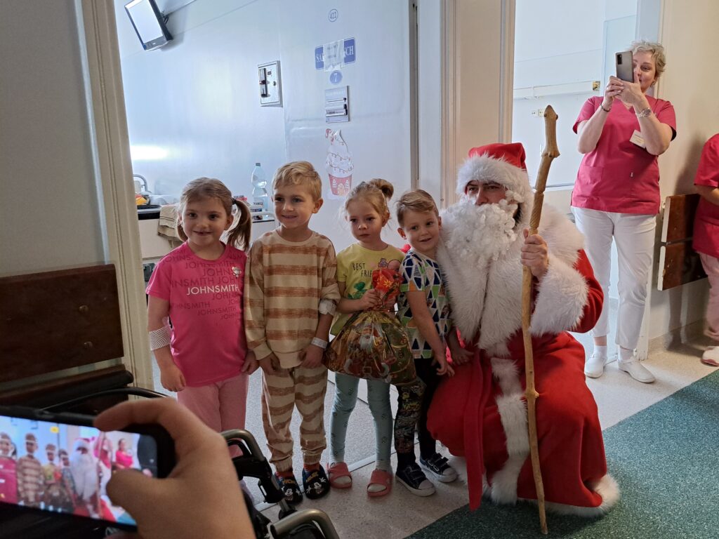 Mikołaj pozuje do zdjęcia z czwórką dzieci: dwoma chłopca i dwiema dziewczynkami, dzieci stoją, Mikołaj przykucnął
