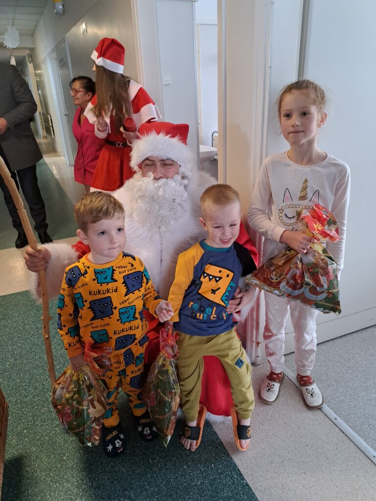 Mikołaj pozuje do zdjęcia z trójką dzieci, dwaj chłopcy siadają mu na kolanach, dziewczynka stoi