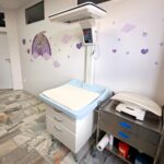 gabinet poradni neonatologicznej, na pierwszym planie stół do badań, na ścianie królik w tęczy w odcieniach fioletu, serduszka i chmurki