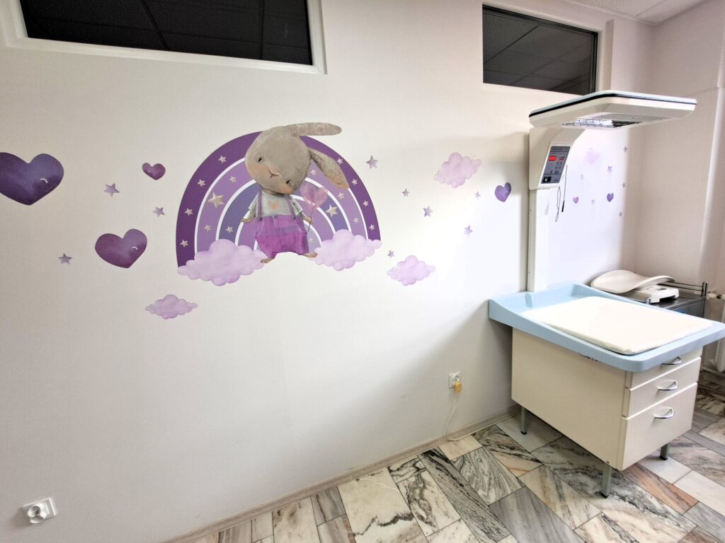 gabinet poradni neonatologicznej, w rogu stół do badań, na ścianie królik w tęczy w odcieniach fioletu, serduszka i chmurki