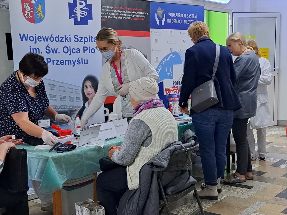 trwają badania poziomu ciśnienia krwi, glukozy i cholesterolu, przy stoliku jedna osoba, dwie pielęgniarki, obok trzy osoby rozmawiają i przeglądają ulotki