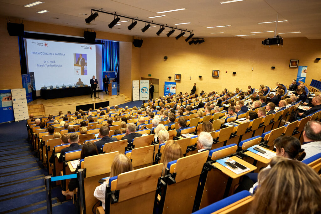 sala konferencyjna, widok z tyłu, rzędy są stopniowane jak w kinie, ludzie siedzą na krzesłach, na scenie przemawia przewodniczący kapituły