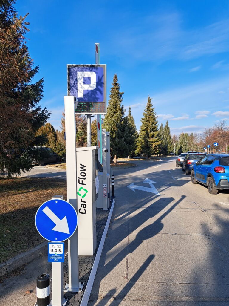 terminal parkingowy z urządzeniem do wydawania biletów, szlabanem i kamerami, przed nimi stoi znak drogowy "nakaz jazdy z prawej strony znaku", a z tyłu znak informacyjny "Parking", po prawej stronie widać zaparkowane samochody