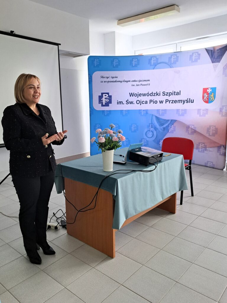 stoi i mówi dyrektorka szpitala, obok stolik z projektorem multimedialnym, za nią biały ekran i ścianka promocyjna