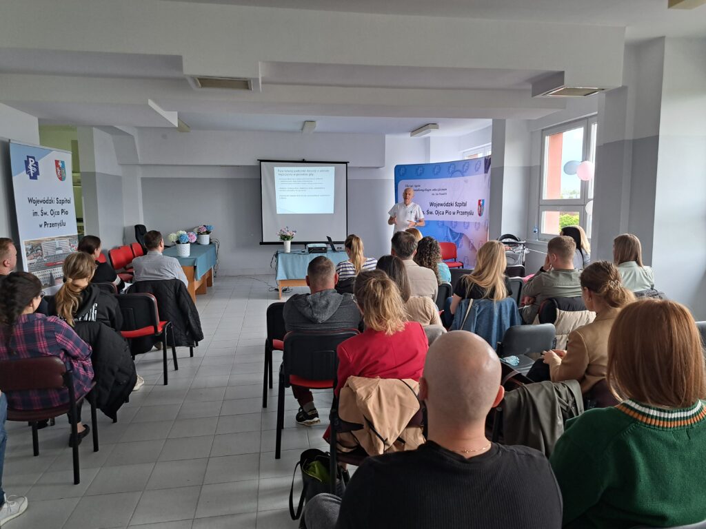 Sala pełna ludzi, obróceni plecami do obiektywu, w tle lekarz prowadzi wykład