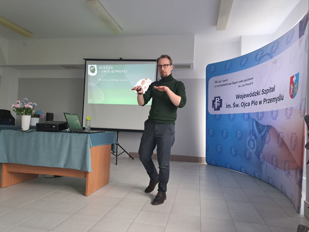 mówi psycholog, obok po prawej stolik z projektorem multimedialnym, za nim biały ekran multimedialny, a po lewej ścianka promocyjna