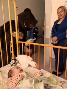 Osoba przebrana za misia na sali szpitalnej, na łóżeczku leży kilkutygodniowe niemowlę, obok dyrektorka spzitala