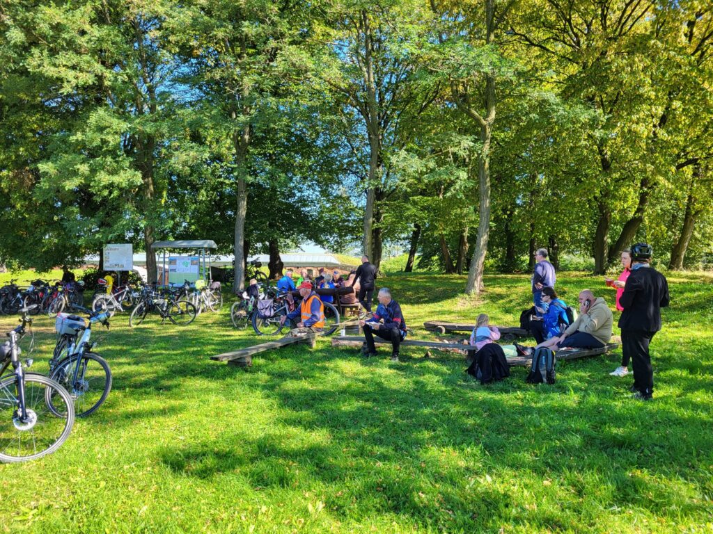 uczestnicy na forcie wśród zieleni, niektórzy siedzą na ławkach, inni stoją, wokół dużo drzew, trawa