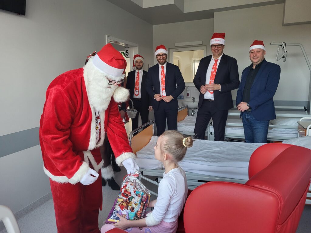 Mikołaj wręcza prezent siedzącej dziewczynce, w tle prezydent, zastępca i zastępca dyrektora