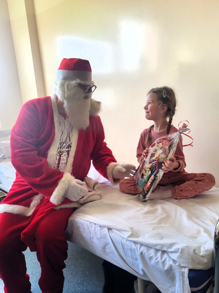 św. Mikołaj siedzi na łóżku obok dziewczynki z prezentem