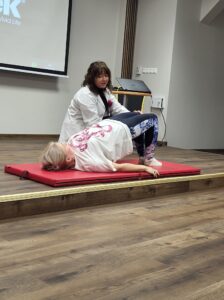 kobieta na materacu wykonuje ćwiczenia, obok pomaga fizjoterapeutka