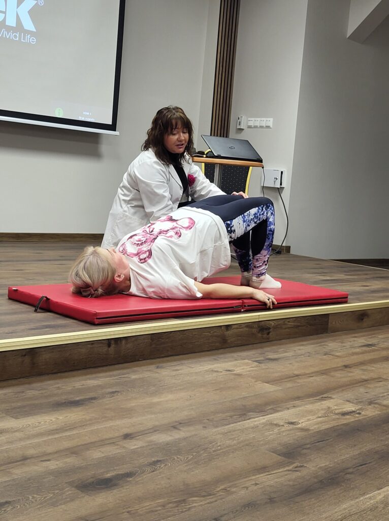 kobieta na materacu wykonuje ćwiczenia, obok pomaga fizjoterapeutka