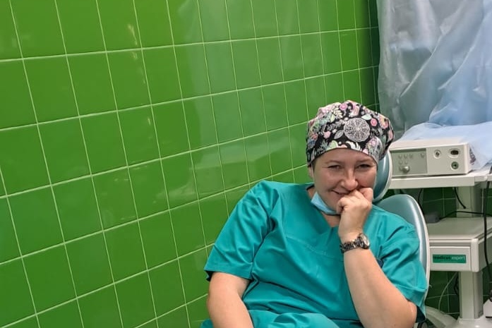 uśmiechnięta lekarka z maską chirurgiczną zsuniętą na brodę siedzi na krześle w pokoju zabiegowym, ręką podpiera głowę