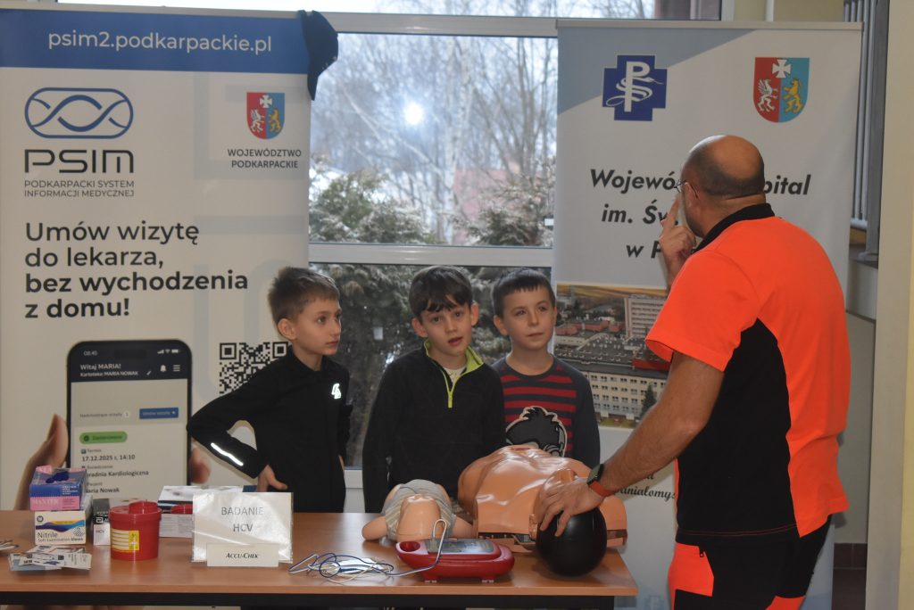 ratownik udziela instrukcji z zakresu pierwszej pomocy, obok stoją trzej chłopcy, na stoliku fantom i akcesoria do nauki