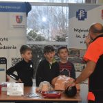 ratownik udziela instrukcji z zakresu pierwszej pomocy, obok stoją trzej chłopcy, na stoliku fantom i akcesoria do nauki