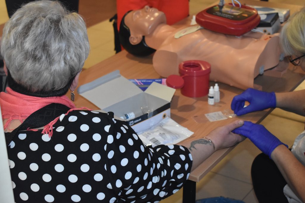 badanie z krwi palca, kobieta siedzi przy stoliku, w tle widać fantom do nauki pierwszej pomocy