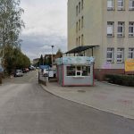 zdjęcie wjazdu na parking, otwarta brama, przy niej budka z pieczywem, a dalej duży budynek przychodni