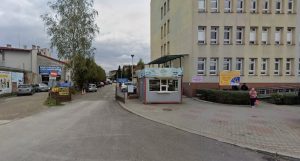 zdjęcie wjazdu na parking, otwarta brama, przy niej budka z pieczywem, a dalej duży budynek przychodni
