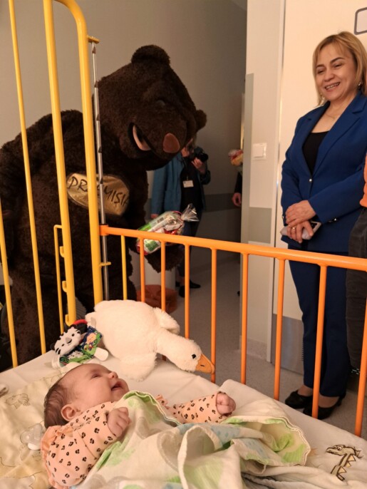 Osoba przebrana za misia na sali szpitalnej, na łóżeczku leży kilkutygodniowe niemowlę, obok dyrektorka spzitala