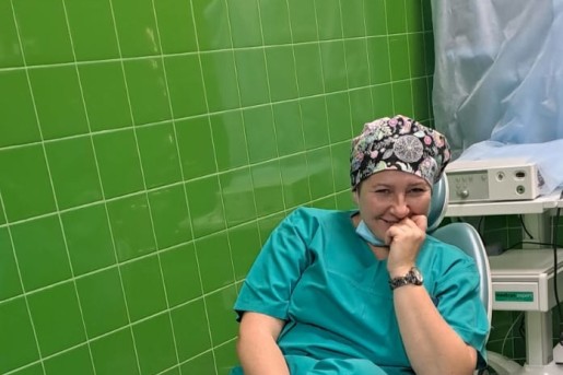 uśmiechnięta lekarka z maską chirurgiczną zsuniętą na brodę siedzi na krześle w pokoju zabiegowym, ręką podpiera głowę