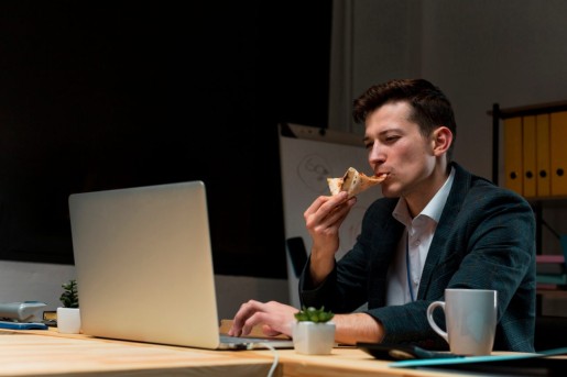 mężczyzna przy laptopie je kawałek pizzy, w pokoju panuje mrok